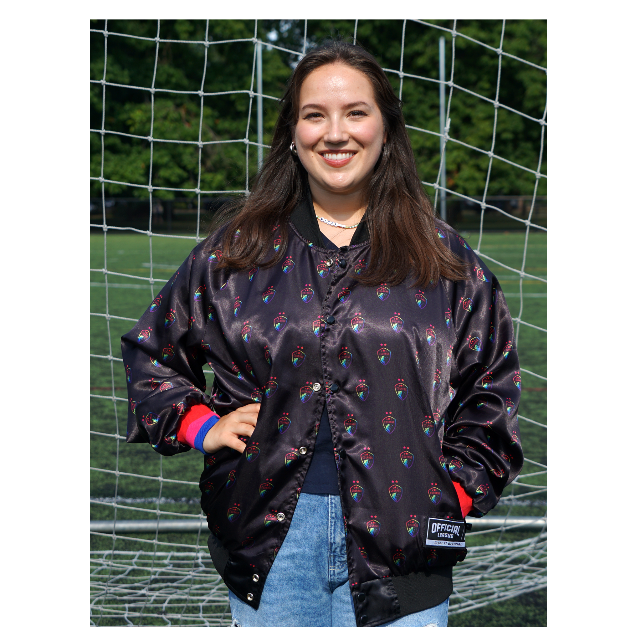 A person wearing a black satin jacket inside out with the pride Courage logo printed all over the jacket, standing next to a soccer goal.