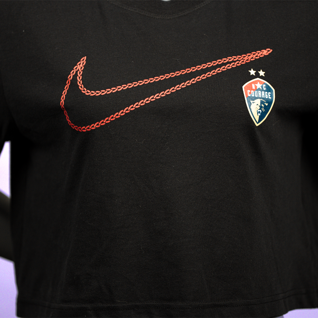 A close up of a black crop top featuring the Nike Swoosh logo in red and the NC Courage logo on the left chest area, displayed on a mannequin.