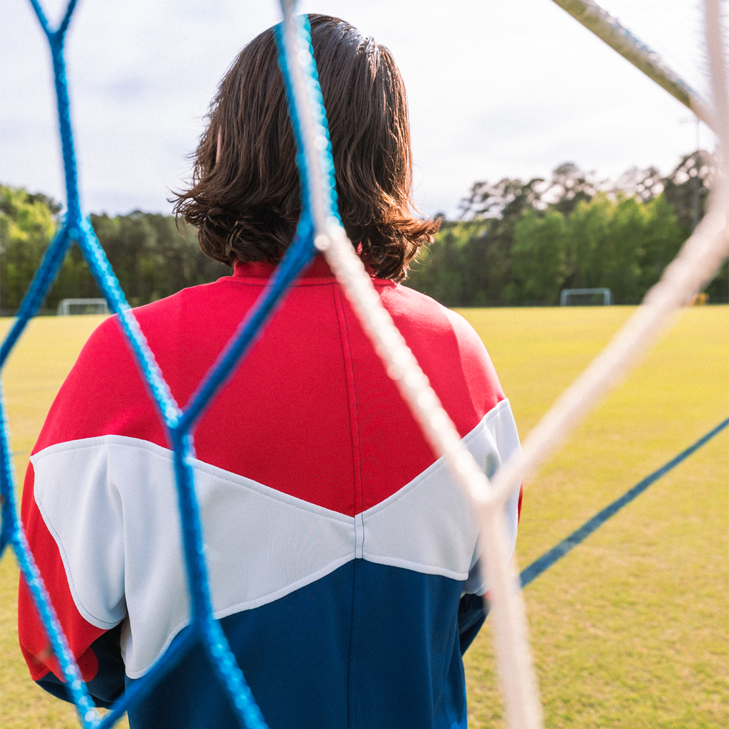 Person wearing a red, white, and blue jacket standing behind a soccer goal on a field.