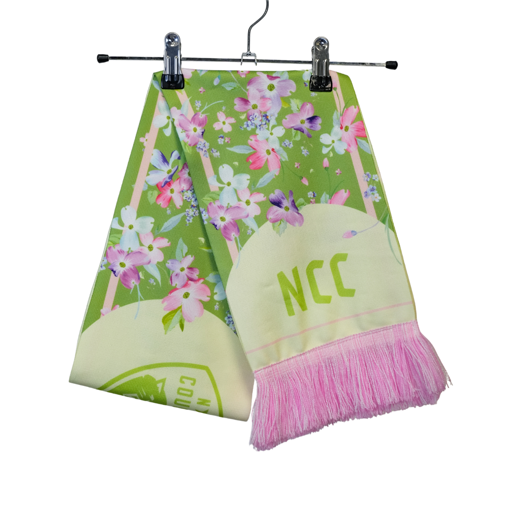 Folded scarf on the hanger. Green background with pink and purple pastel flowers. Pink fringe and "NCC" in green