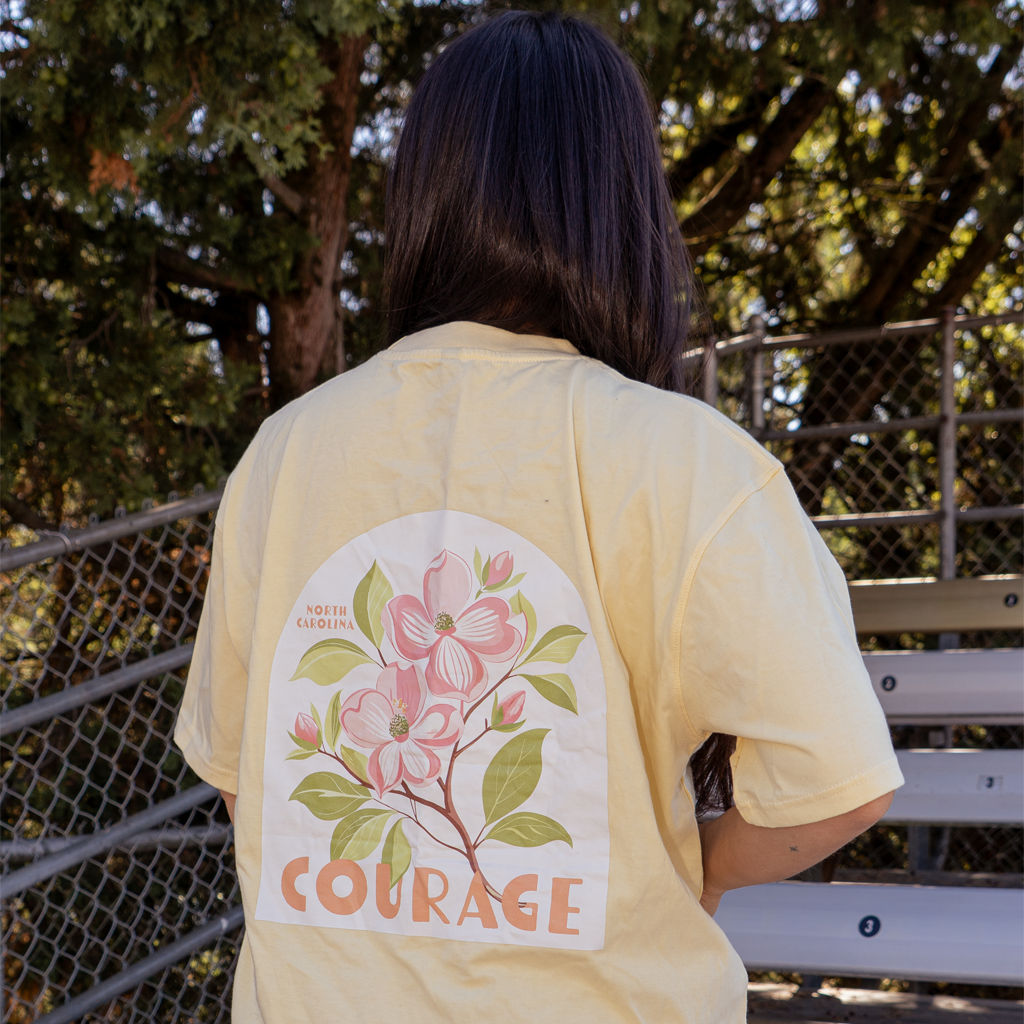 Model posing showing the back of yellow t-shirt with a white arch shaped filled with pink dogwood flowers and Courage underneath