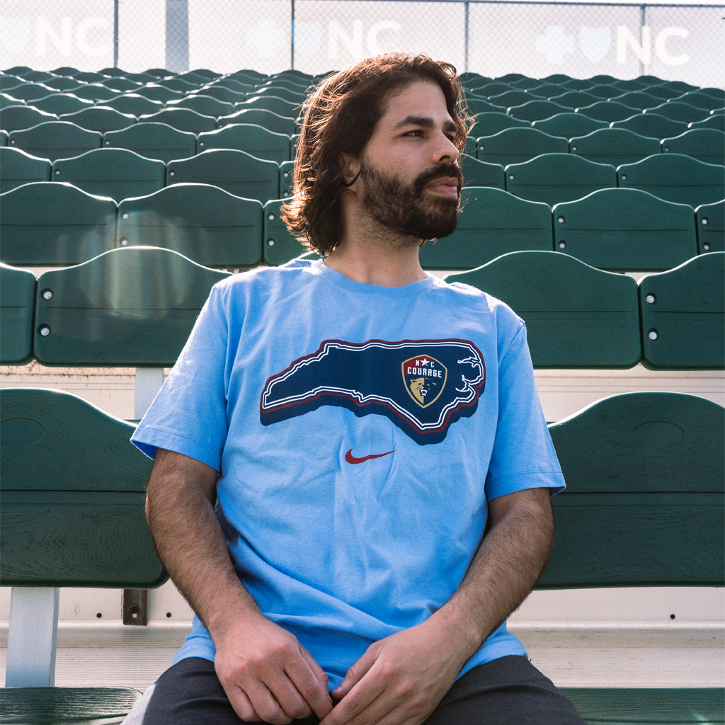 Man sitting on a green bench wearing a blue t-shirt with the North Carolina Courage logo, in a stadium.