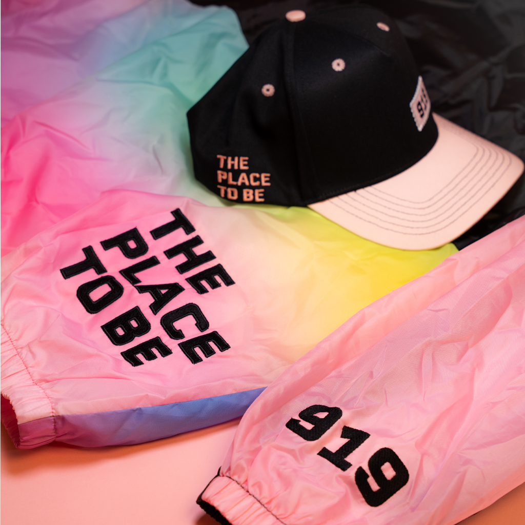 Colorful jacket with 'The Place to Be' text and a cap on a pink background
