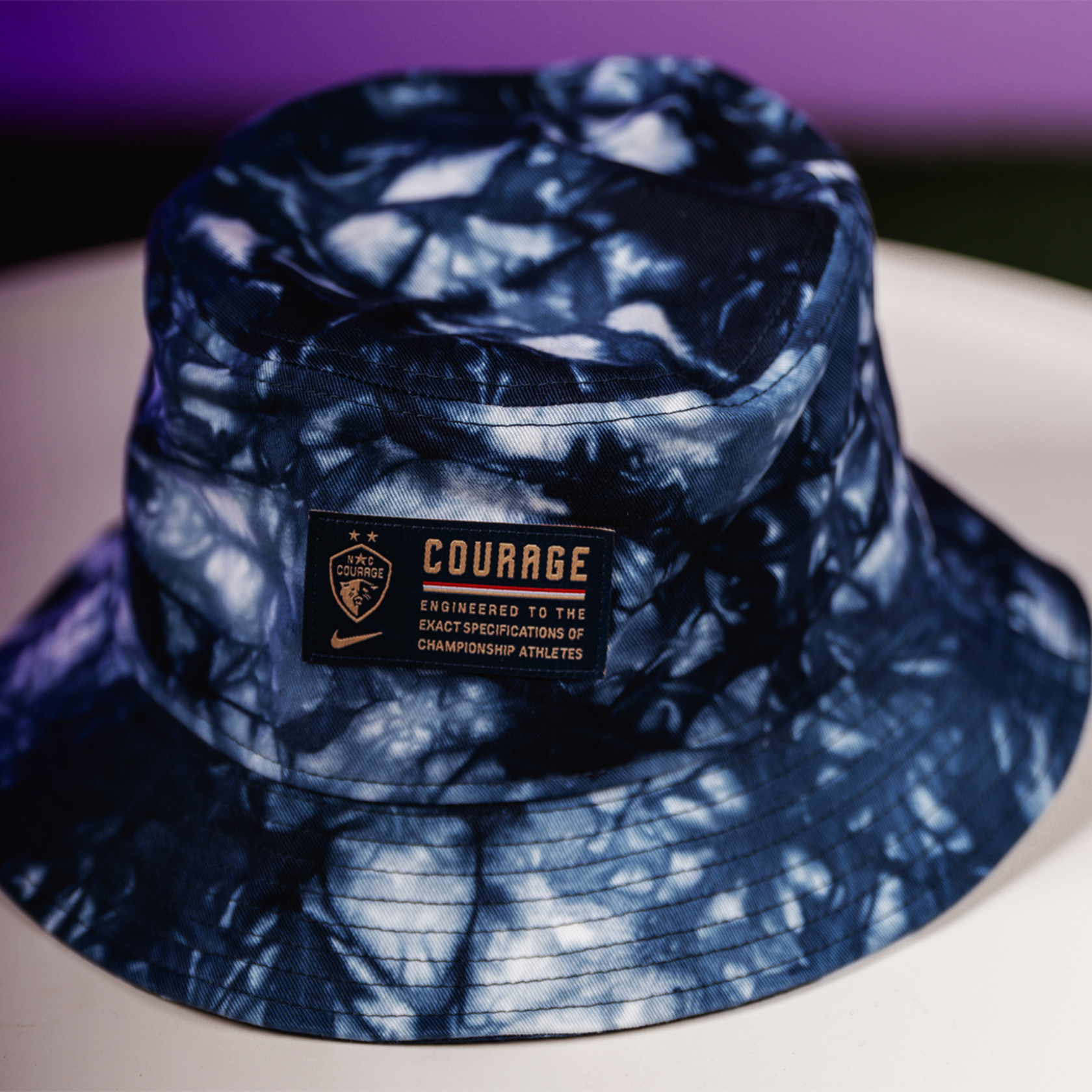 A tie-dye bucket hat with the NC Courage crest and a label that reads ' COURAGE TRUSTED TO THE CHAMPIONSHIP ATHLETES'.