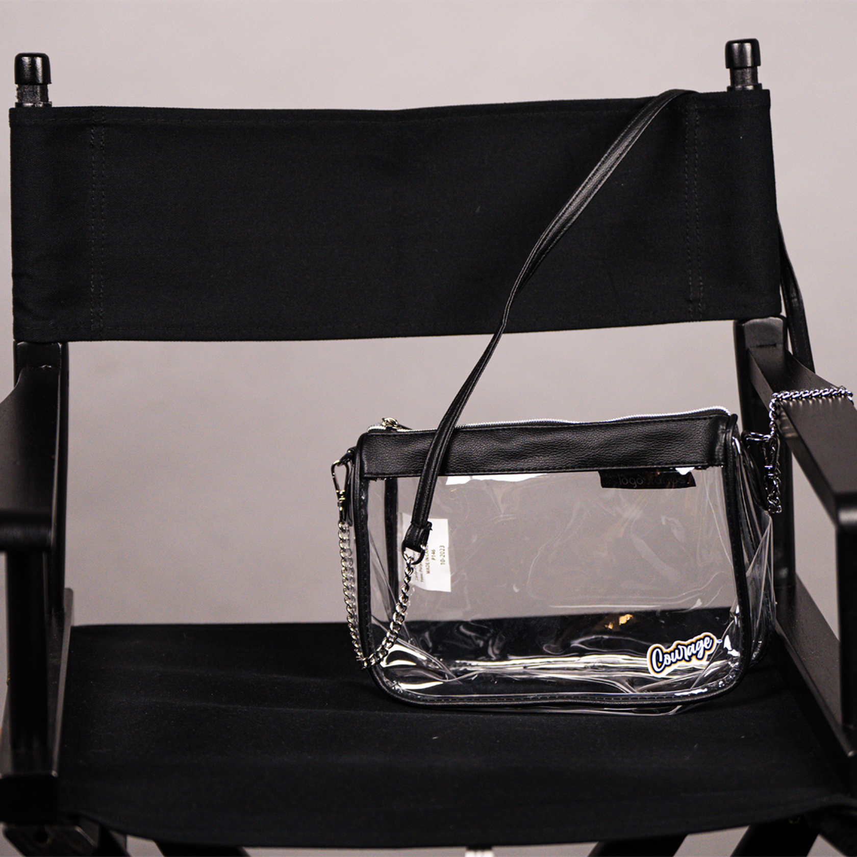 A clear plastic shoulder bag with a black strap and black trim, featuring the NC Courage logo, displayed on a black chair.