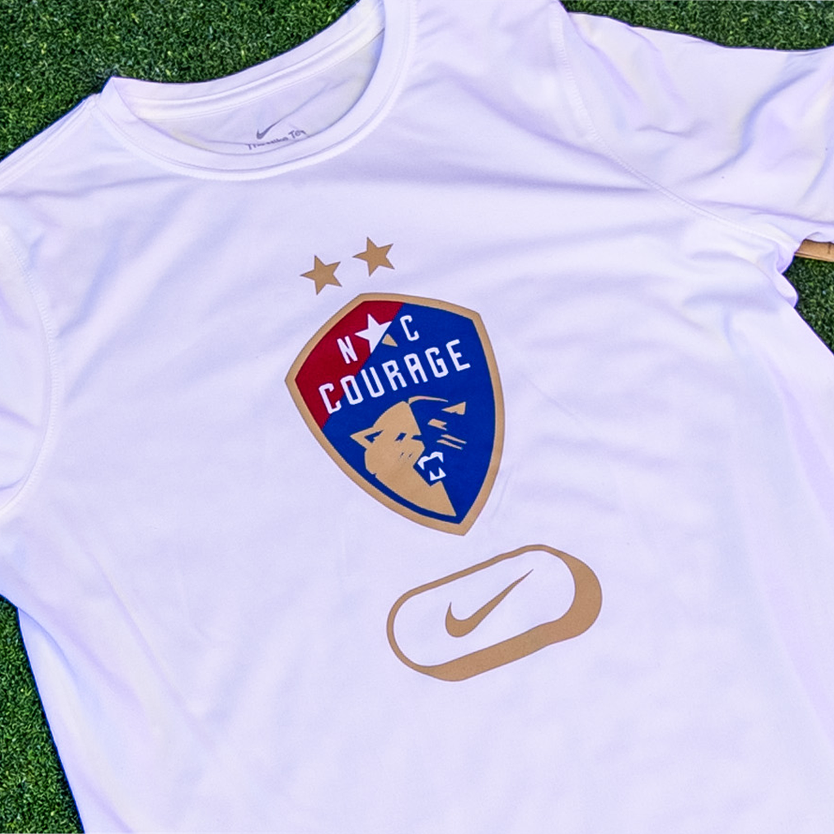 A zoom in of a white tee with a Courage Logo on the front and a gold nike logo.