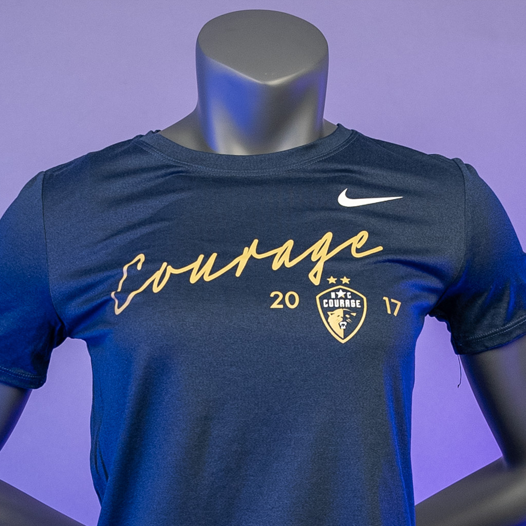 A close up of a navy blue t-shirt with the word 'COURAGE' printed in gold above a graphic emblem, displayed on a mannequin against a purple.