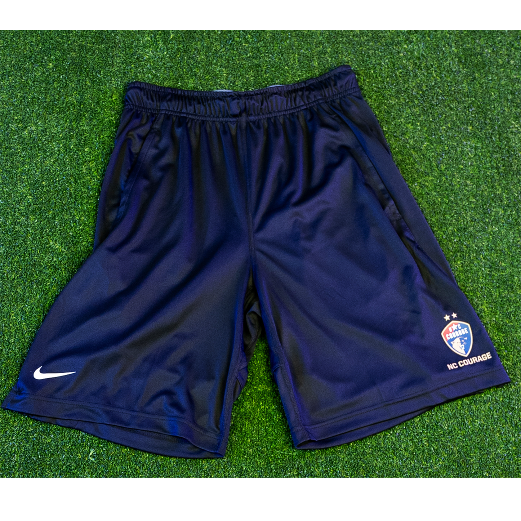 A pair of men's navy sports shorts with a logo on the left leg, displayed on a green background.