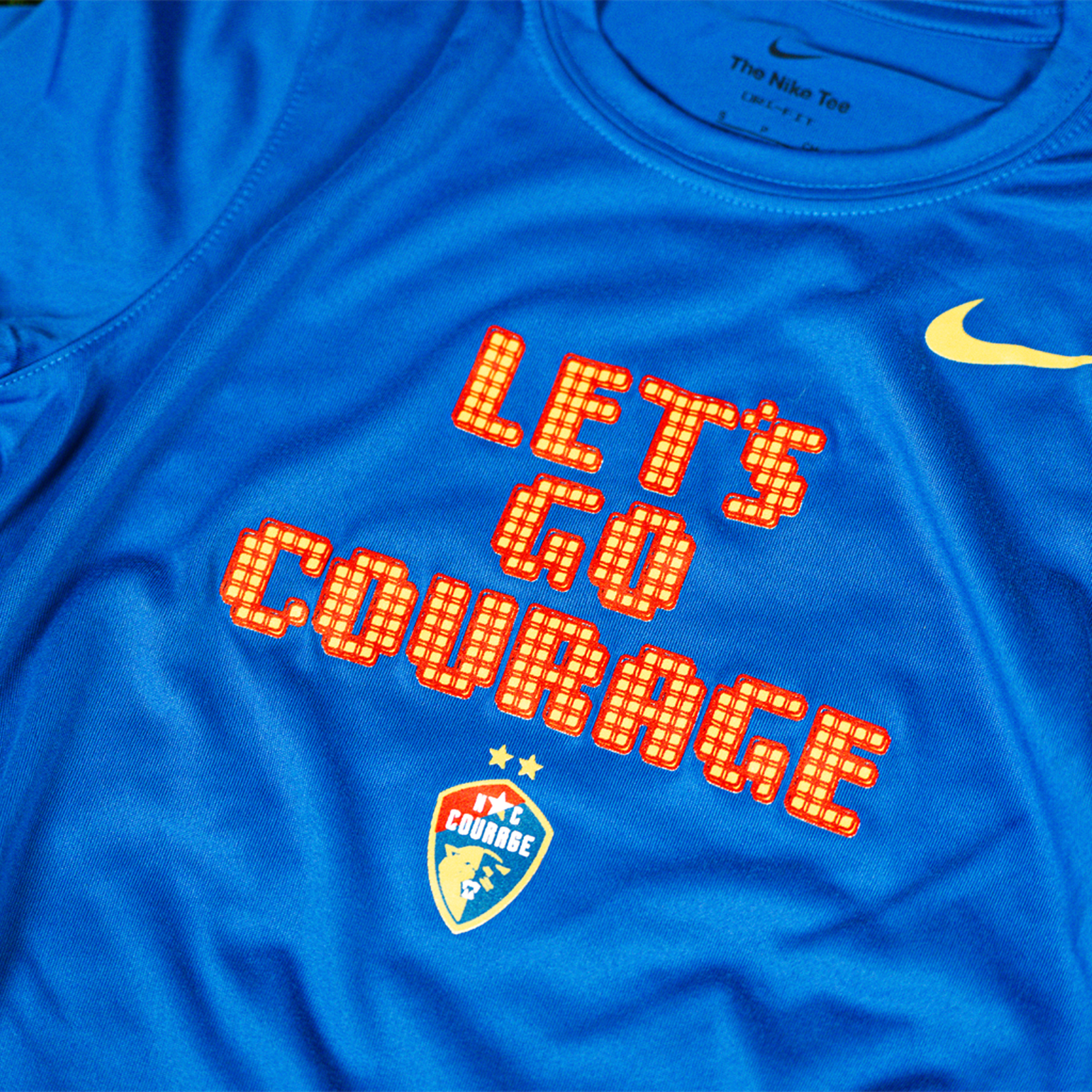 A close up of a blue NC Courage Youth Pixel Dri-Fit Tee with the text 'LET'S GO COURAGE' and the NC Courage logo on it, placed on a grassy surface.
