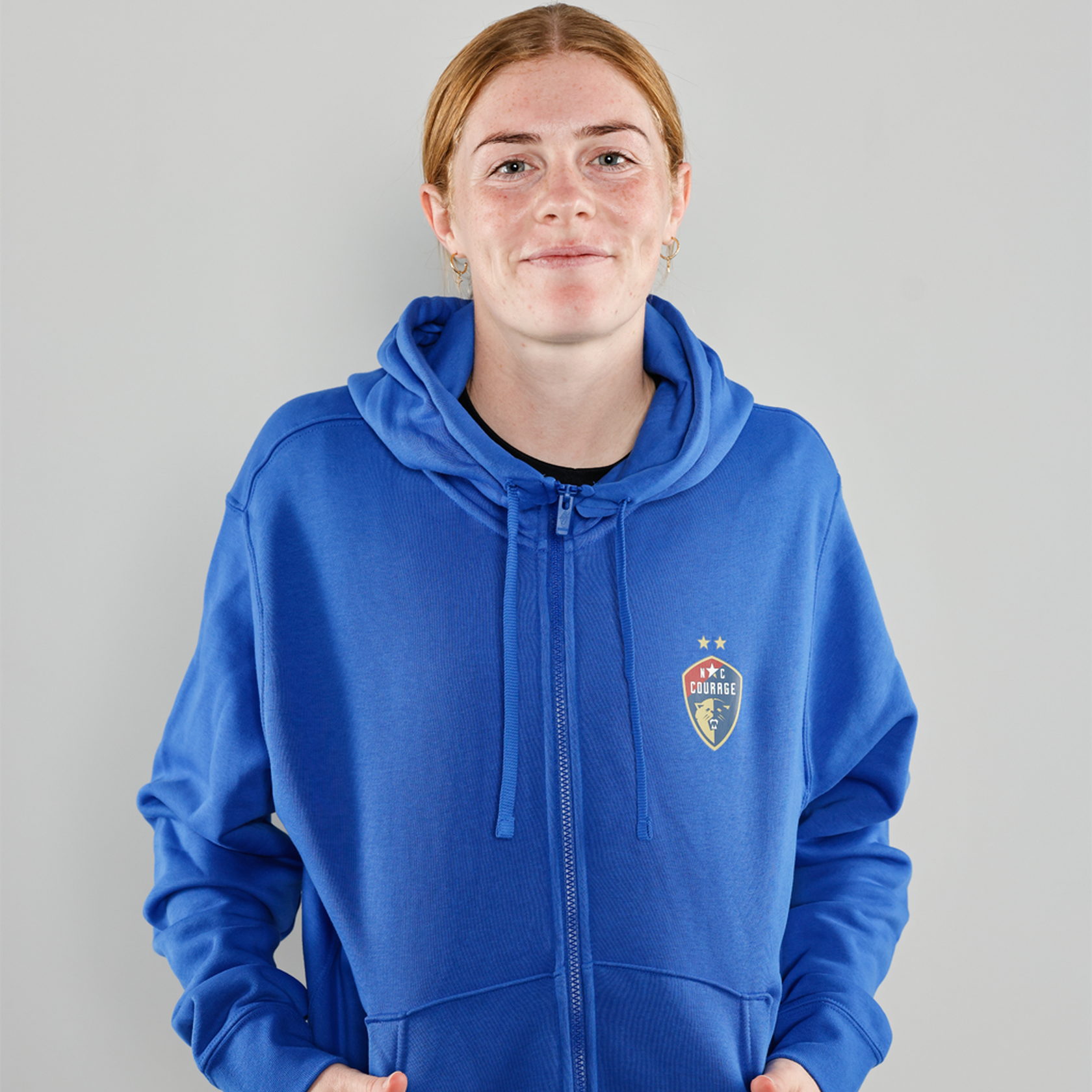 Cortnee Vine models a royal blue full zip hoodie with the NC Courage logo displayed on the upper left front side.