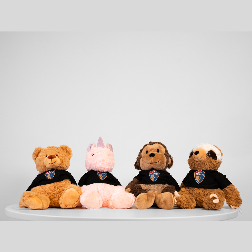 Four plush toy animals, including a bear, a pink pig, a wolf, and a dog, each wearing a black shirt with a logo, seated in a row on a white surface against a neutral background.