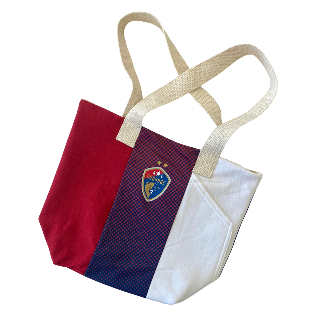 A canvas tote bag with horizontal blue, red, and white panels, featuring the North Carolina Courage logo on the front.