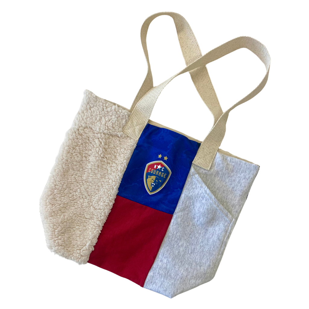 A canvas tote bag with horizontal blue, red, and white panels, featuring the North Carolina Courage logo on the front.