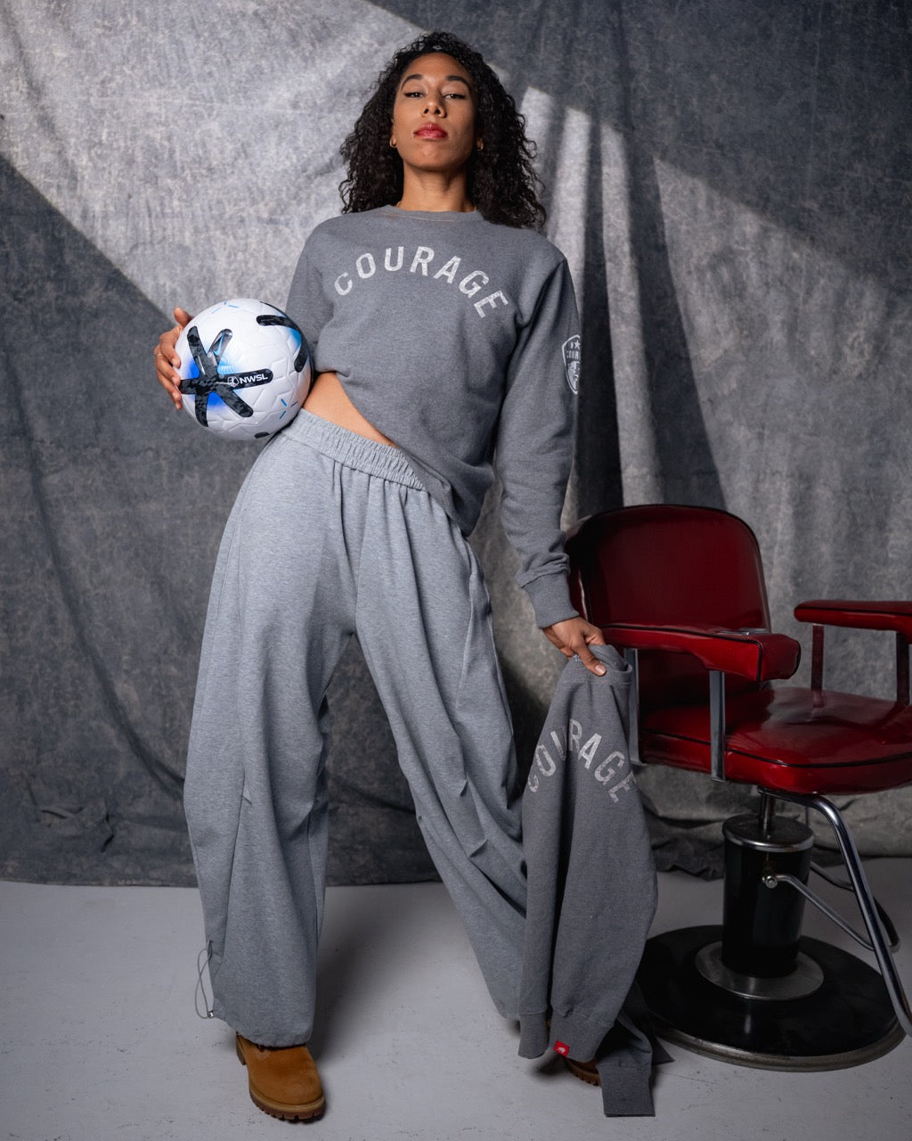 Person wearing a 'COURAGE' sweatshirt and holding a soccer ball in a studio setting with a red chair.