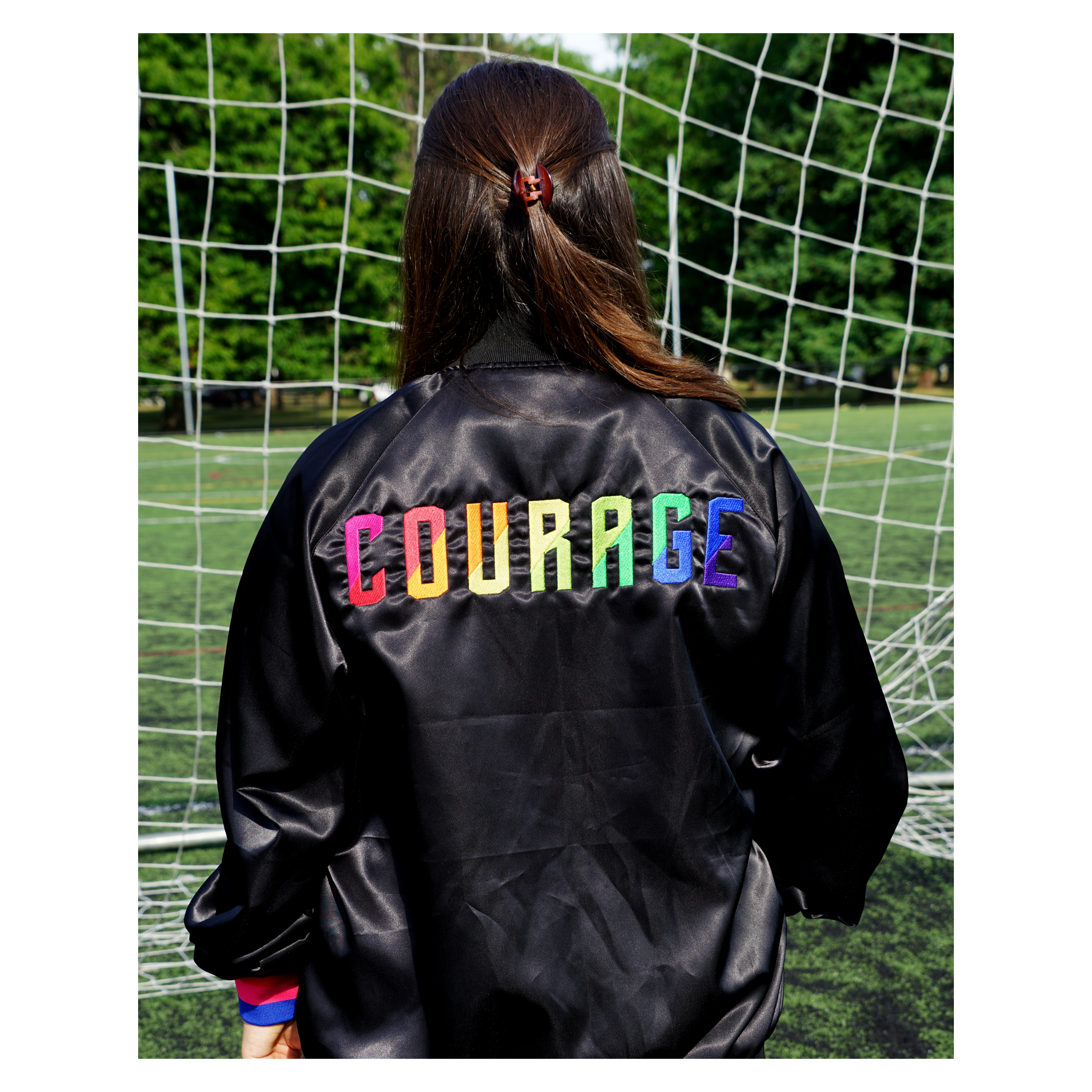 A person wearing a black satin jacket with the Courage embroidered Courage text in the color of the rainbow, standing next to a soccer goal.