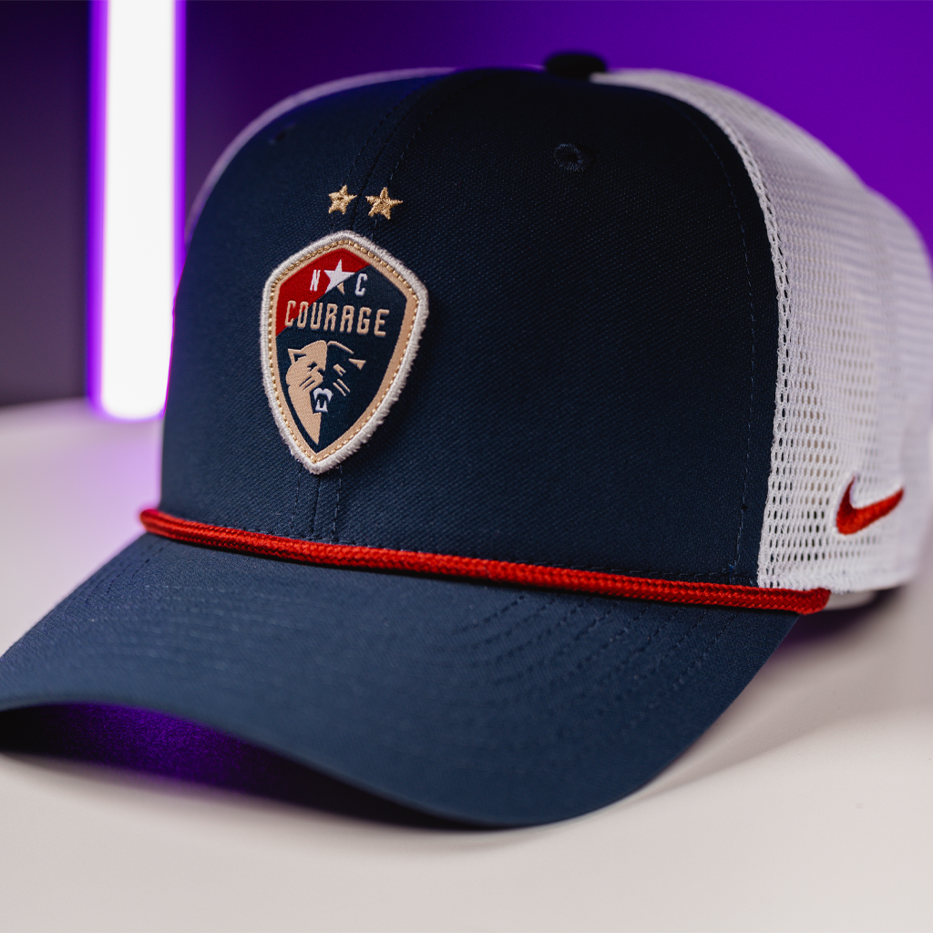 A side view of a trucker hat with a rope detail on the brim and a ' Courage' sports team logo on the front against a purple background.
