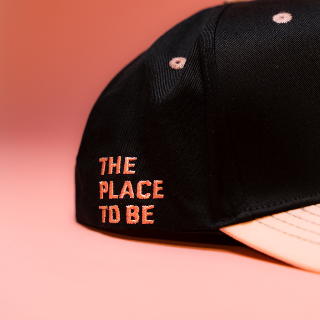 Black cap with 'THE PLACE TO BE' text on a pink background