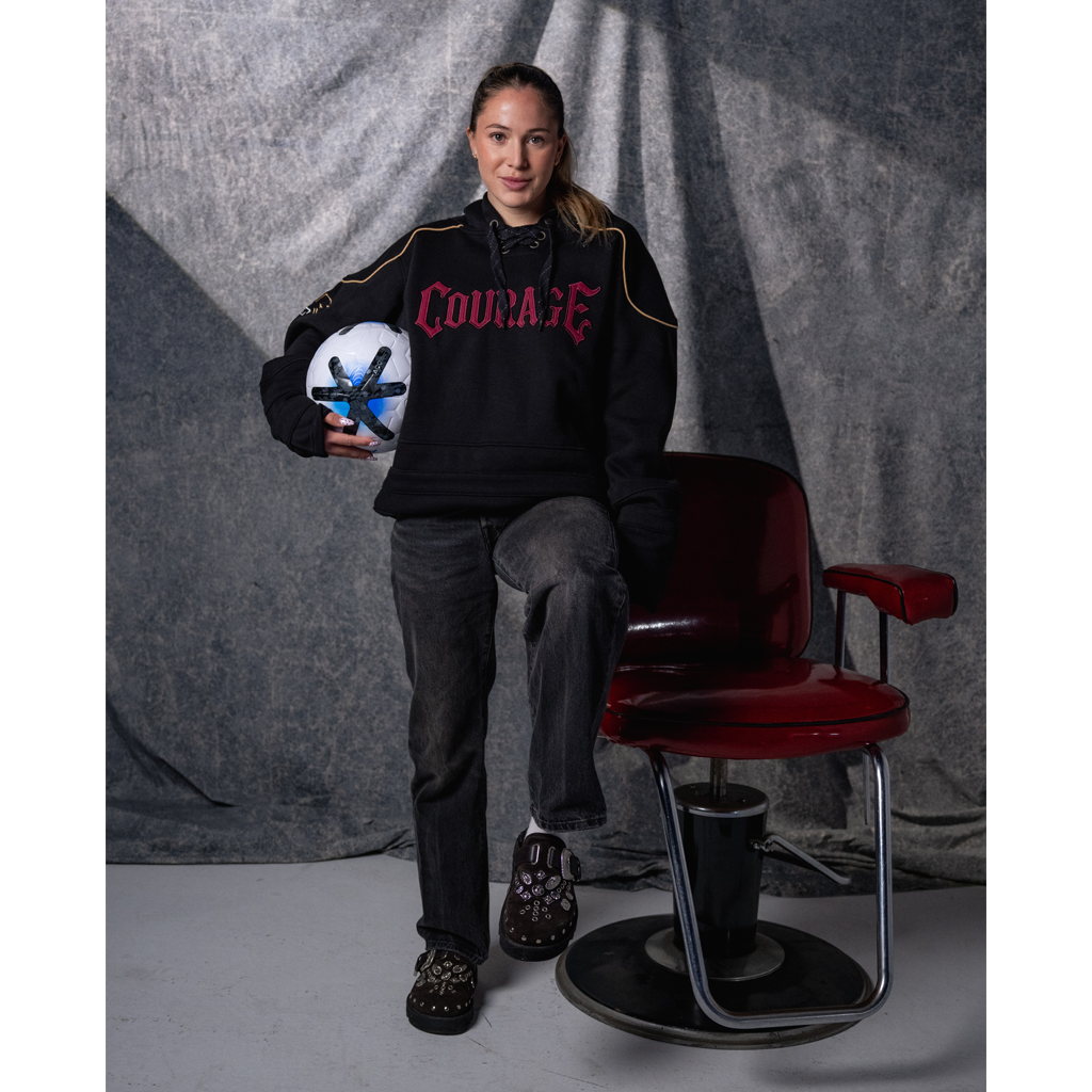 Person wearing a black sweatshirt with 'COURAGE' text, holding a ball, sitting on a red chair against a gray curtain background.