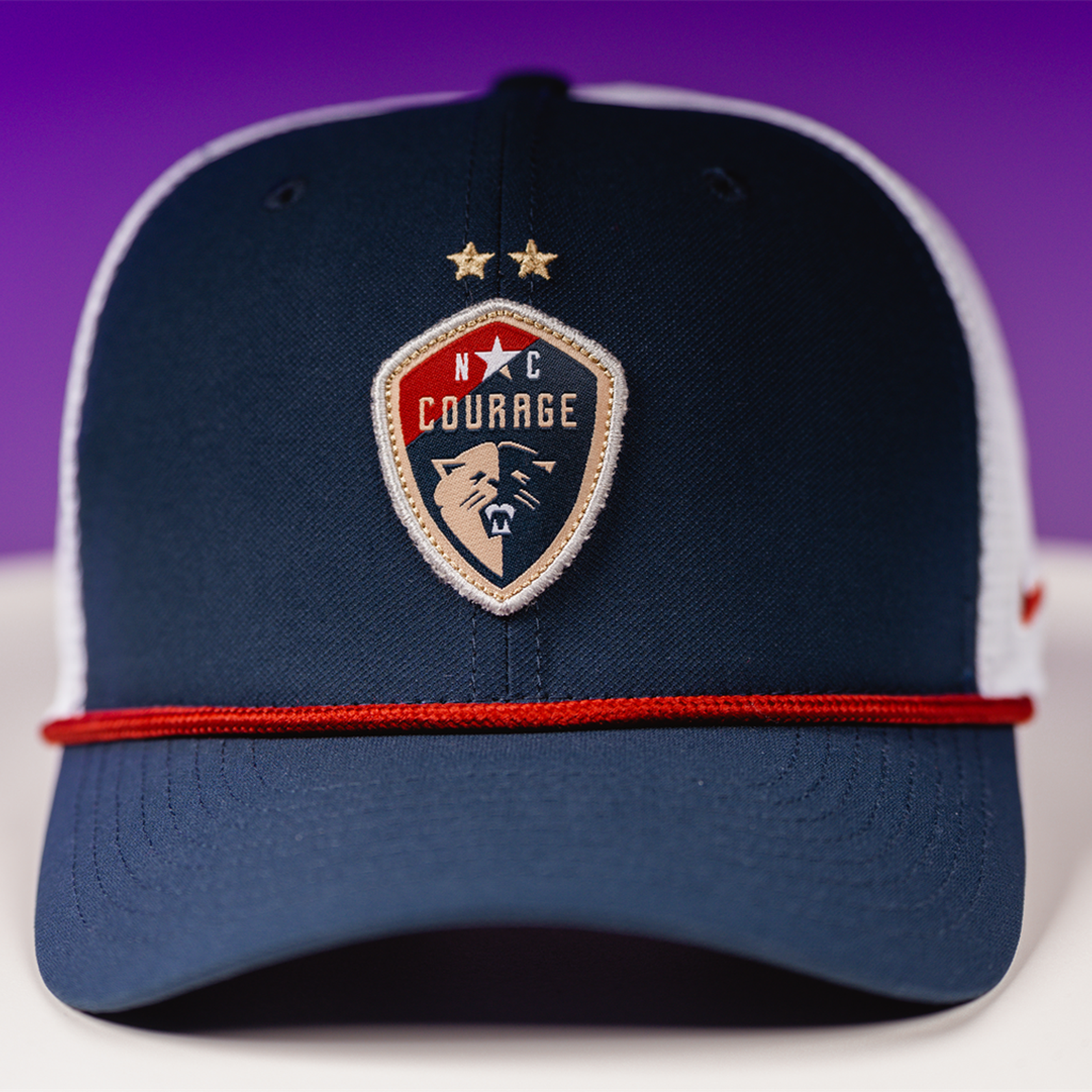 A trucker hat with a rope detail on the brim and a ' Courage' sports team logo on the front against a purple background.