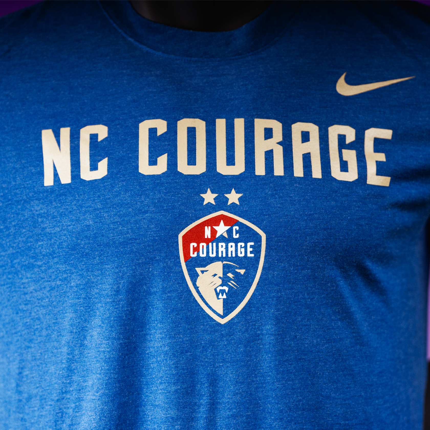 A close up of a blue t-shirt with 'NC COURAGE' written in white and the NC Courage logo in white and red.