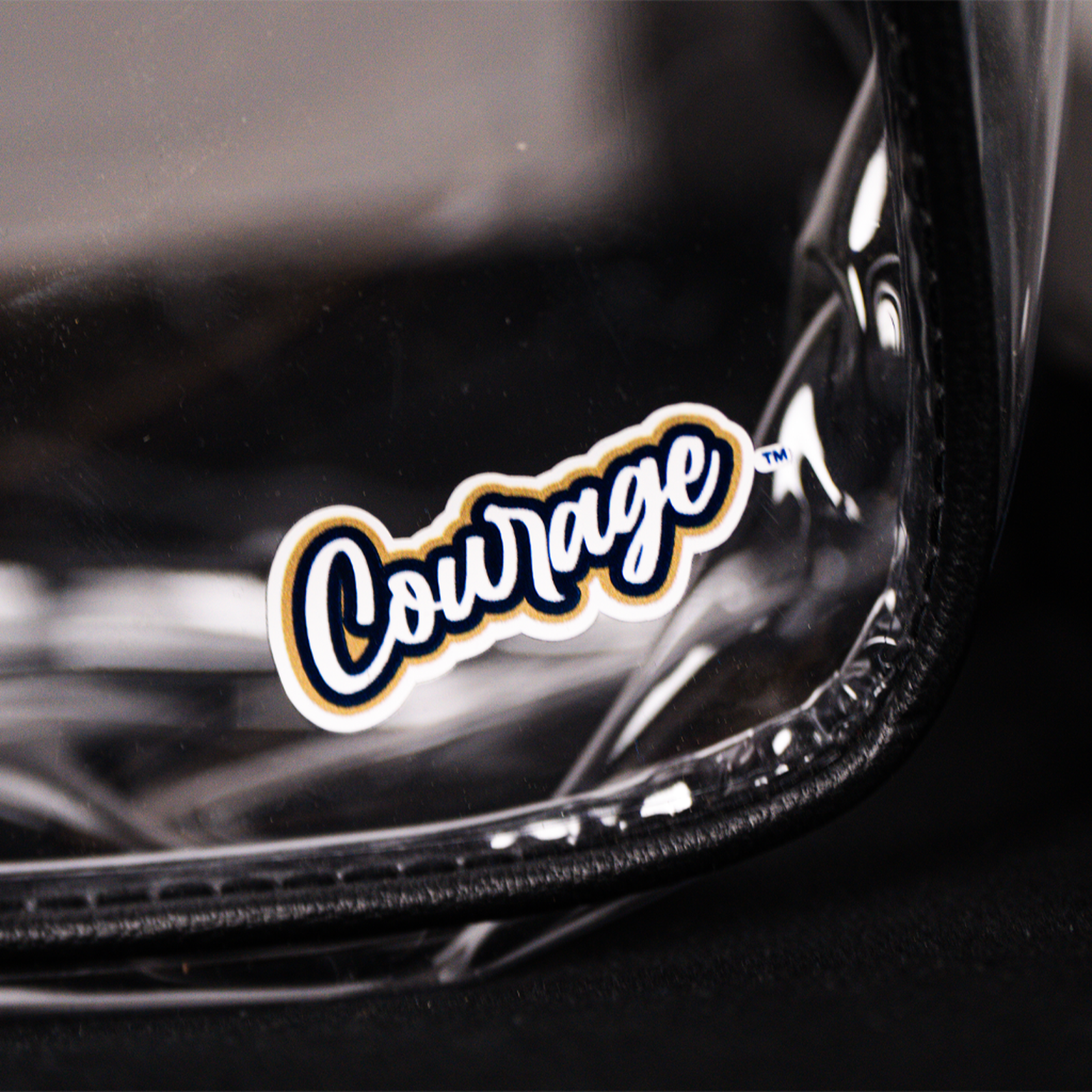 A close up of a clear plastic shoulder bag with a black strap and black trim, featuring the NC Courage logo, displayed on a black chair.