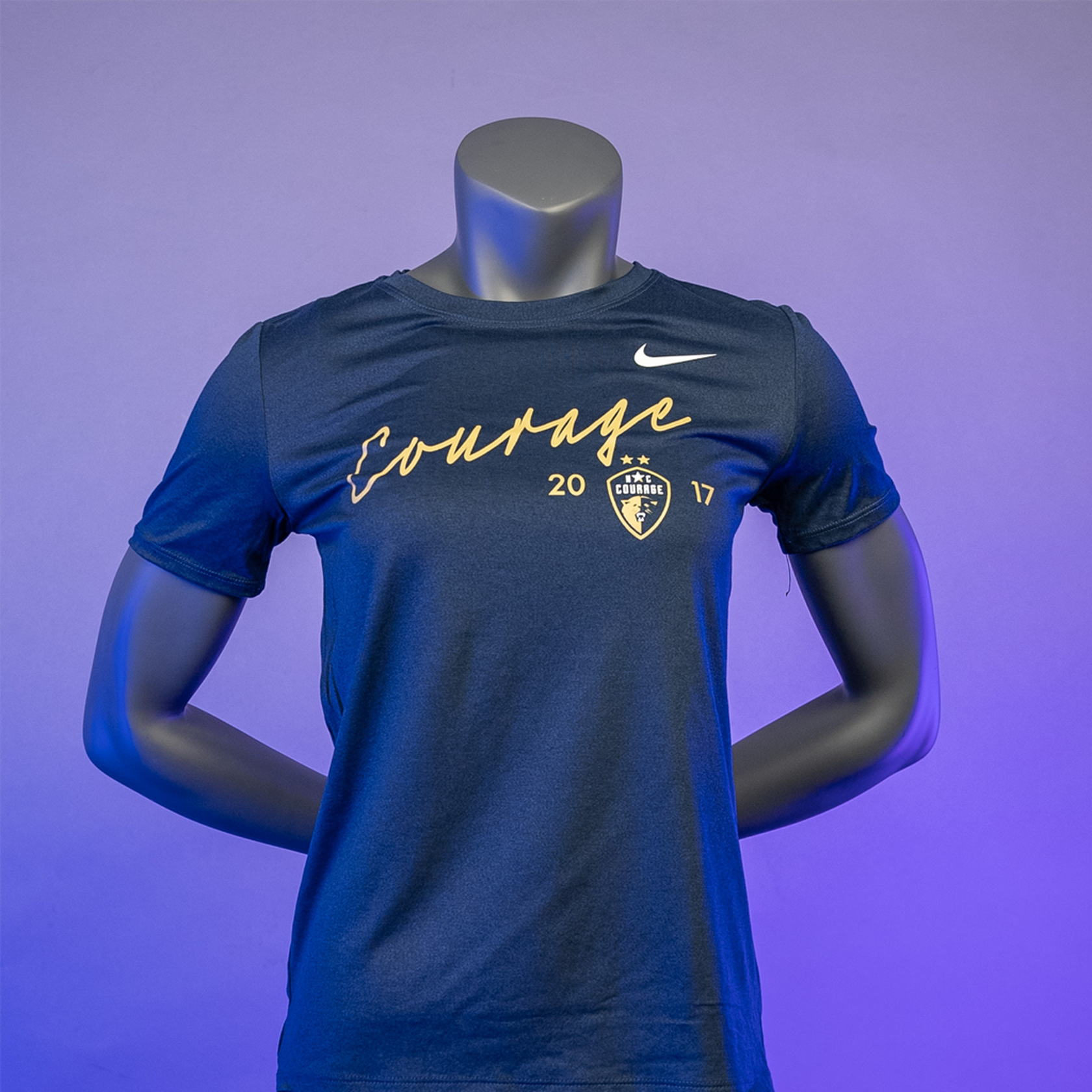 A navy blue t-shirt with the word 'COURAGE' printed in gold above a graphic emblem, displayed on a mannequin against a purple background.