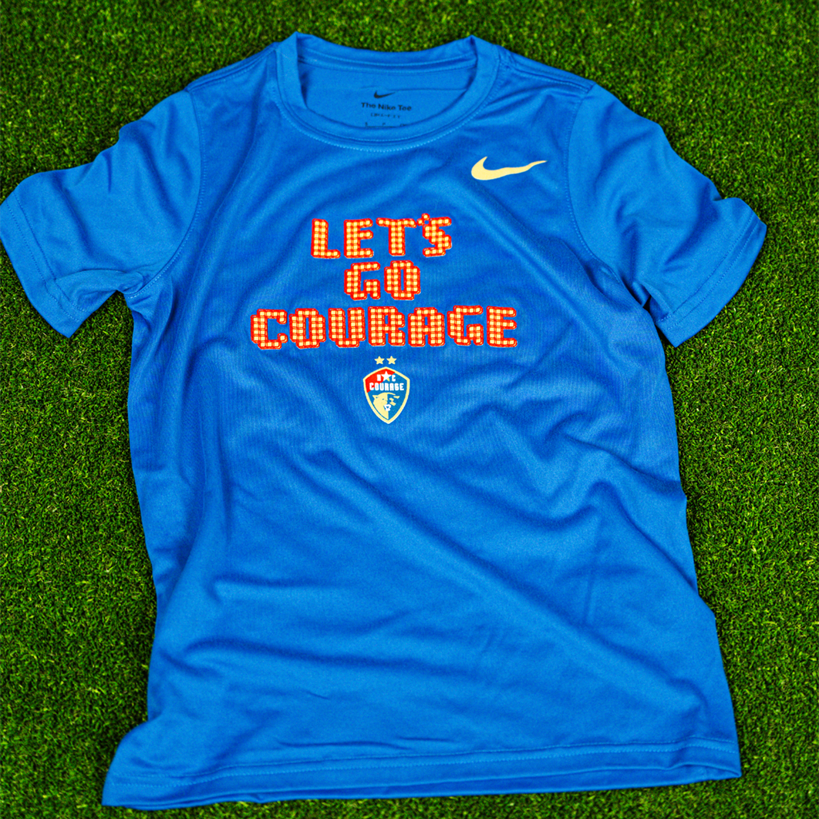 A blue NC Courage Youth Pixel Dri-Fit Tee with the text 'LET'S GO COURAGE' and the NC Courage logo on it, placed on a grassy surface.