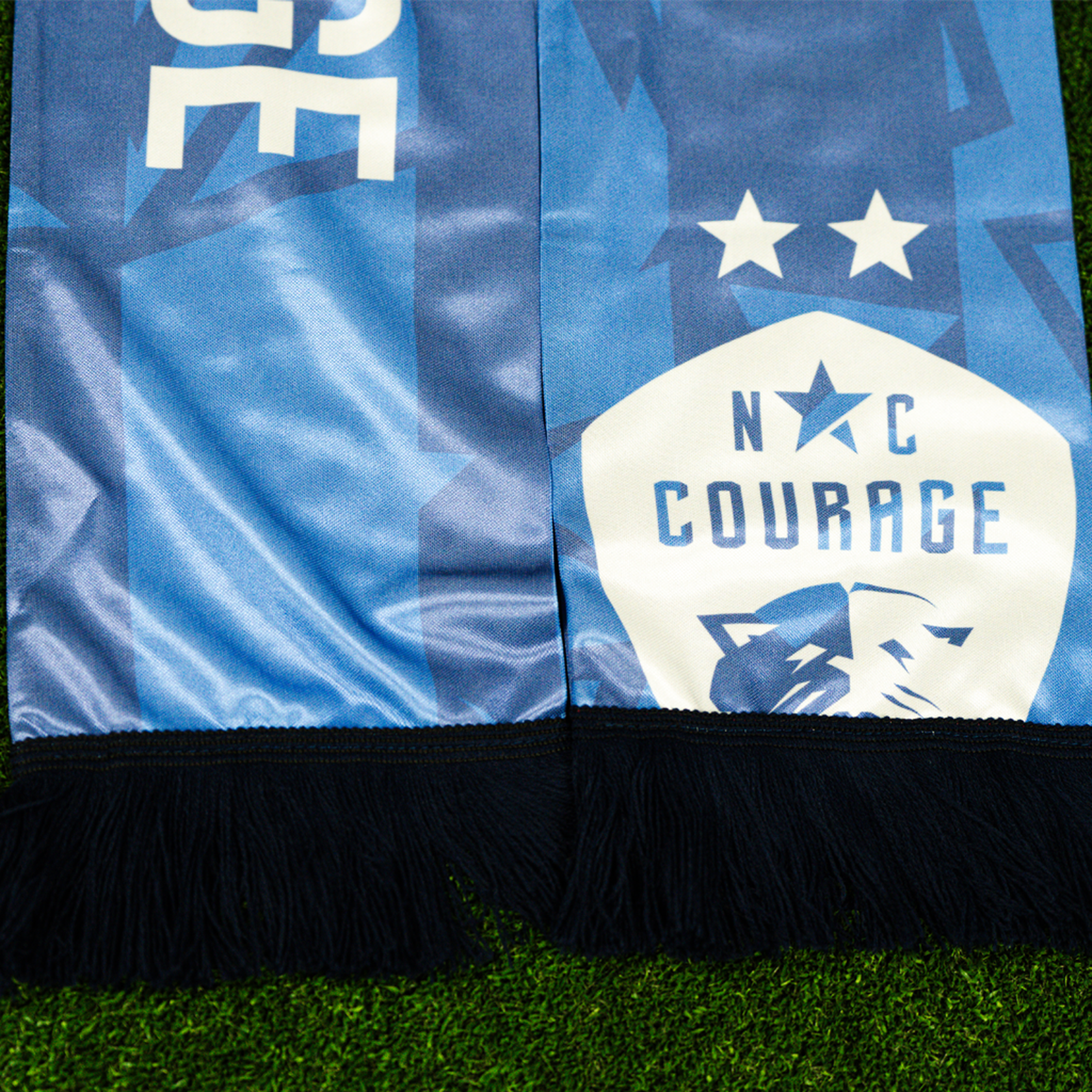 A zoom in of a purple scarf with the text 'ALL COURAGE' and the NC Courage logo, displayed on a green grass-like surface.