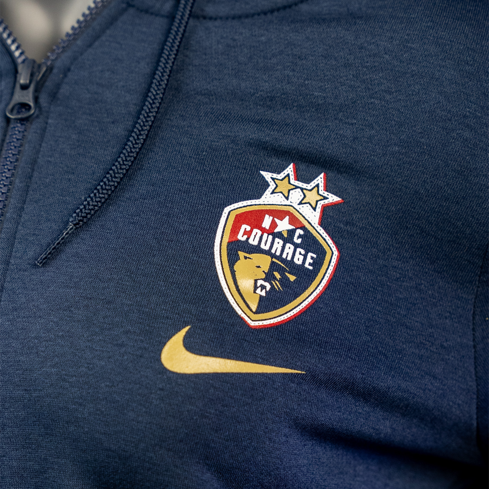 A close up of a navy blue women's full zip jacket with a hood, featuring a logo on the chest.