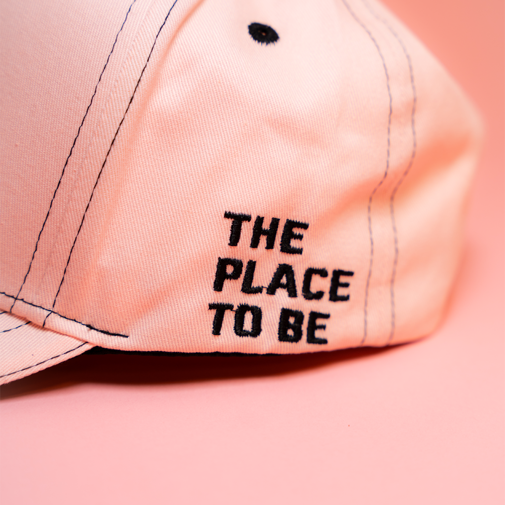 Pink cap with 'THE PLACE TO BE' text on a pink background