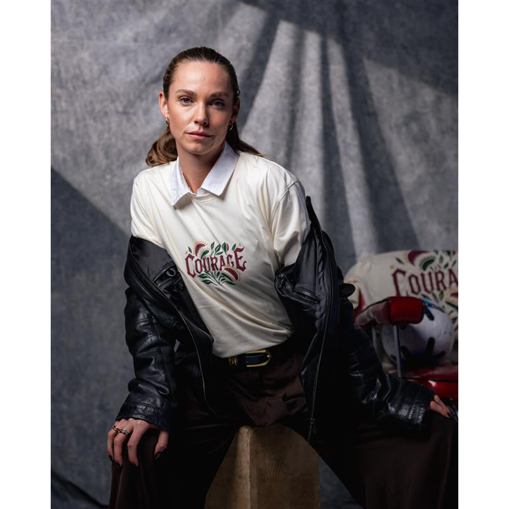 Woman wearing a white shirt with 'Courage' printed on it, sitting next to a motorcycle.